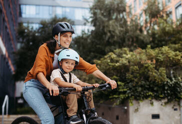 Mutter, die ihren Sohn auf einem sicheren Kinderfahrradträger oder -sitz trägt, beide mit Helm. Mutter, die mit einem kleinen Kind auf dem Fahrrad durch die Stadt fährt. - HPIF32205