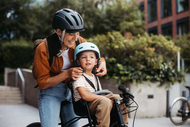 Mutter, die ihrem Sohn den Fahrradhelm auf den Kopf setzt und ihn auf dem Kinderfahrradträger oder -sitz trägt. Mutter, die mit einem kleinen Kind mit dem Fahrrad durch die Stadt fährt. - HPIF32206