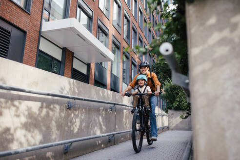 Mother carring her son on a secure child bike carrier or seat, both wearing helmets. Mom commuting with a young child through the city on a bicycle. - HPIF32207