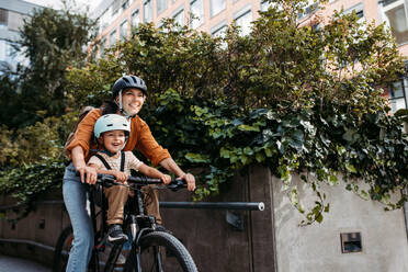 Mutter, die ihren Sohn auf einem sicheren Kinderfahrradträger oder -sitz trägt, beide mit Helm. Mutter, die mit einem kleinen Kind auf dem Fahrrad durch die Stadt fährt. - HPIF32208