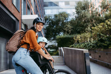 Mother carring her son on a secure child bike carrier or seat, both wearing helmets. Mom commuting with a young child through the city on a bicycle. - HPIF32209