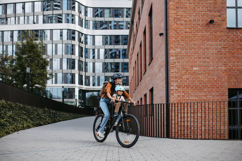 Mother carring her son on a secure child bike carrier or seat, both wearing helmets. Mom commuting with a young child through the city on a bicycle. - HPIF32211