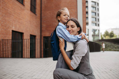 Eine berufstätige Mutter umarmt ihre Tochter vor dem Schulgebäude und macht sich auf den Weg zur Arbeit. Konzept der Work-Life-Balance für Frauen. - HPIF32222