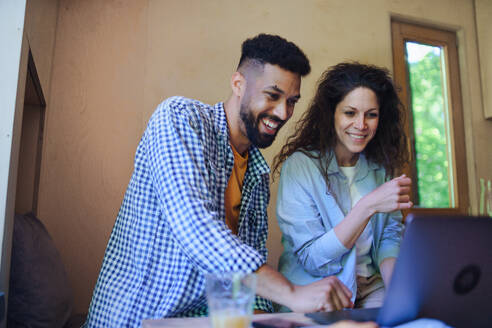 A happy couple with laptop sitting, resting and talking indoors in a tree house, remote office concept. - HPIF32232