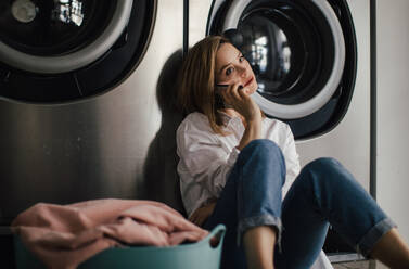 Young woman calling with smartphone, sitting in a laundry room. - HPIF32259