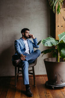 Portrait of a young businessman sitting near window in modern office full of plants. Male corporate leader, executive, manager making phone call. - HPIF32370