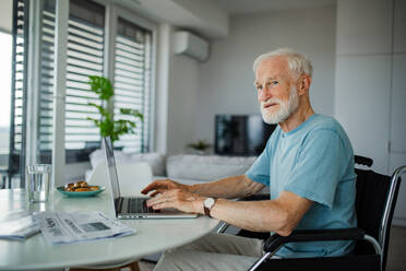 Älterer Mann im Rollstuhl, der mit einem Laptop online einkauft. Älterer Mann, der digitale Technologien nutzt und in seiner Küche an einem Notebook arbeitet. Konzept von Senioren und digitalen Fähigkeiten. - HPIF32385