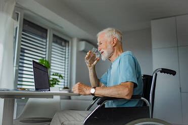 Älterer Mann im Rollstuhl, der mit einem Laptop online einkauft. Älterer Mann, der digitale Technologien nutzt und in seiner Küche an einem Notebook arbeitet. Konzept von Senioren und digitalen Fähigkeiten. - HPIF32386
