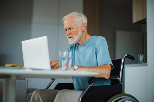 Senior man in a wheelchair working from home during retirement. Elderly man using digital technologies, working on a laptop. Concept of seniors and digital skills. - HPIF32387