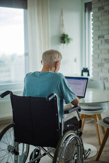 Älterer Mann in einem Rollstuhl, der ein Dokument auf einem Laptop schreibt. Älterer Mann, der digitale Technologien nutzt und in seiner Küche an einem Notebook arbeitet. Konzept der Senioren und digitalen Fähigkeiten. - HPIF32388