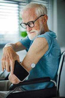 Diabetic senior man checking blood glucose level at home using continuous glucose monitor. Elderly man connecting his CGM with smarphone to see blood sugar levels in real time. - HPIF32393