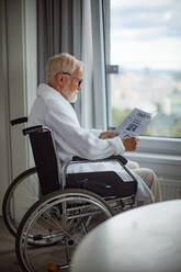 Elderly man in a wheelchair reading the newspaper in his robe in the morning. Concept of loneliness and dependence of retired people. - HPIF32397
