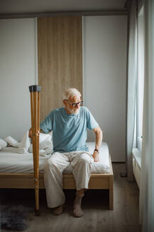 Portrait of an elderly man with crutches trying to stand up from bed. - HPIF32404