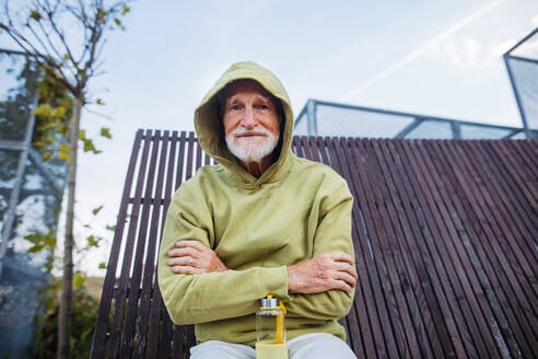 Portrait of cool elderly man in a hoodie with the hood on his head and arms crossed over his chest, during exercising outdoors. - HPIF32410