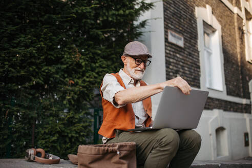 Senior man sitting on street curb working on his laptop outdoors. Portrait of elderly man using digital technologies, working with notebook. Concept of seniors and digital skills. - HPIF32440