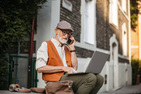 Senior man sitting on street curb working on his laptop outdoors. Portrait of elderly man using digital technologies, working on a notebook. Concept of seniors and digital skills. - HPIF32441