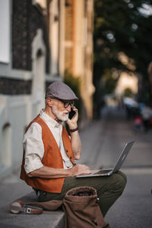 Älterer Mann sitzt auf der Straße und arbeitet im Freien an seinem Laptop. Porträt eines älteren Mannes, der digitale Technologien nutzt und mit Notebook und Smartphone arbeitet. Konzept von Senioren und digitalen Fähigkeiten. - HPIF32442