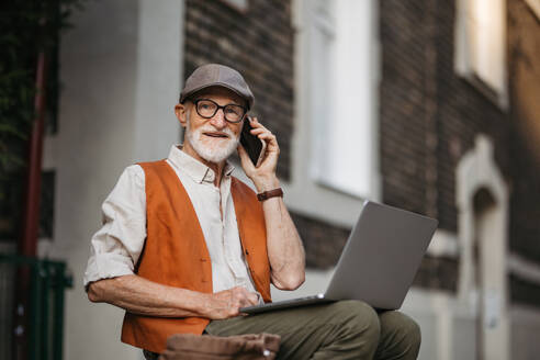 Älterer Mann sitzt auf der Straße und arbeitet im Freien an seinem Laptop. Porträt eines älteren Mannes, der digitale Technologien nutzt und mit Notebook und Smartphone arbeitet. Konzept von Senioren und digitalen Fähigkeiten. - HPIF32443