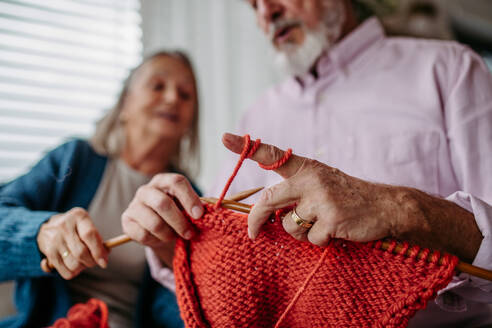 Senior couple knitting together in the living room. An elderly man is learning to knit. - HPIF32500