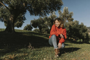 Glückliche Frau hockt in einem Olivenfeld an einem sonnigen Tag - DMGF01137