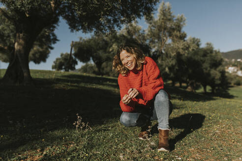 Frau hockt und lacht in einem Olivenfeld an einem sonnigen Tag - DMGF01138