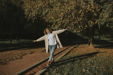 Happy woman balancing on curb of footpath at autumn park - DMGF01147