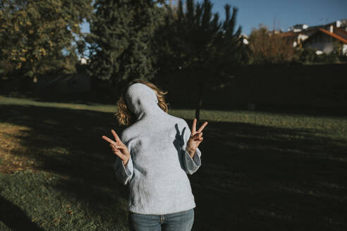 Playful woman covering face with hooded sweater and gesturing peace sign at autumn park - DMGF01154