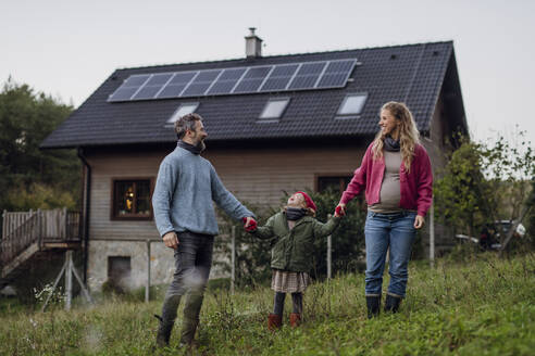 Glückliche Eltern, die ihre Tochter im Gras vor dem Haus an den Händen halten - HAPF03549