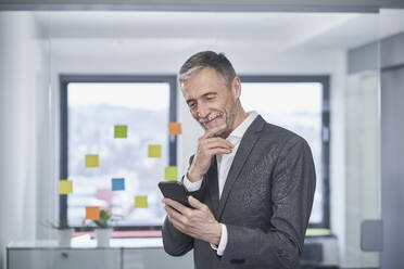 Lächelnder Geschäftsmann mit Hand am Kinn, der ein Smartphone im Büro benutzt - RORF03601