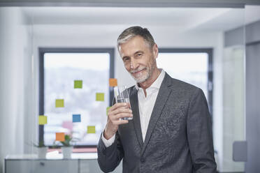 Lächelnder Geschäftsmann mit einem Glas Wasser im Büro stehend - RORF03611