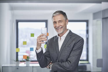 Glücklicher Geschäftsmann mit einem Glas Wasser im Büro stehend - RORF03612