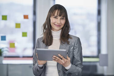 Smiling mature businesswoman standing with tablet PC in office - RORF03622