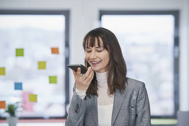 Lächelnde reife Geschäftsfrau, die eine Sprachnachricht auf einem Mobiltelefon im Büro sendet - RORF03625