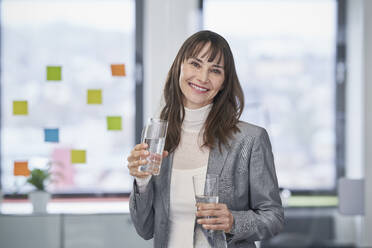 Happy mature businesswoman standing with glass of water and jug - RORF03629