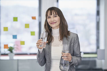 Smiling mature businesswoman holding glass of water and jug in office - RORF03630