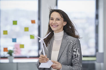 Happy mature businesswoman holding wind turbine model in office - RORF03631