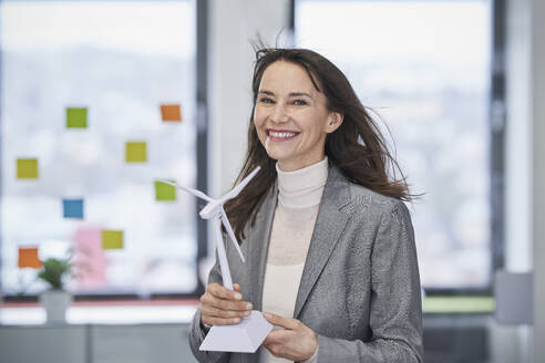 Happy mature businesswoman holding wind turbine model in office - RORF03631