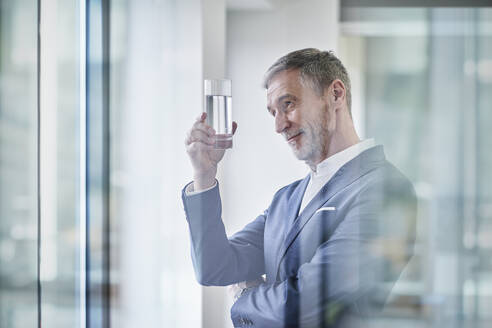 Lächelnder Geschäftsmann, der ein Glas Wasser im Büro untersucht - RORF03636