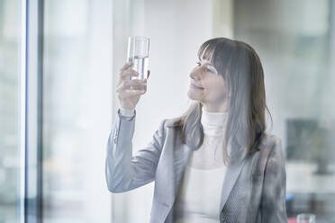 Lächelnde reife Geschäftsfrau, die ein Glas Wasser im Büro untersucht - RORF03638