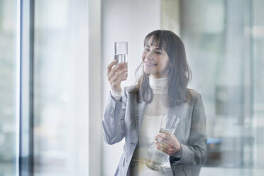 Lächelnde Geschäftsfrau, die ein Glas Wasser untersucht und einen Krug im Büro hält - RORF03640