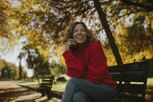 Happy woman sitting and laughing on bench in autumn park - DMGF01167