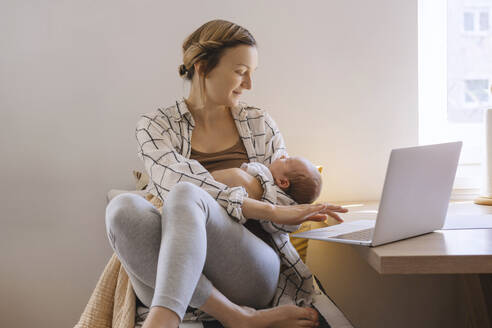 Lächelnde Mutter mit neugeborenem Sohn in den Armen sitzt zu Hause neben dem Laptop - NDEF01479
