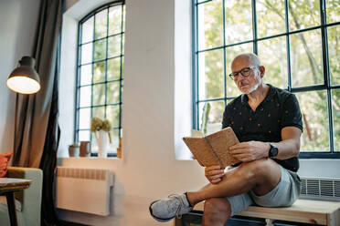 Senior man in community space reading book sitting on bench - HAPF03550