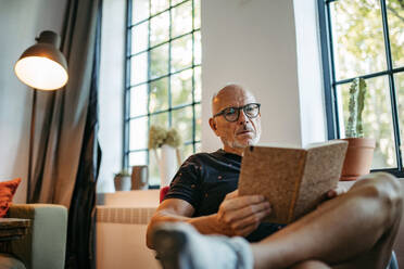 Senior man in community space reading book sitting on bench - HAPF03551