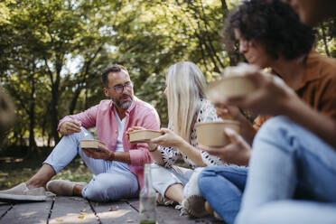 Freiwillige im Gemeinschaftsprojekt essen gemeinsam im Gemeinschaftsgarten, auf dem Boden sitzend - HAPF03623