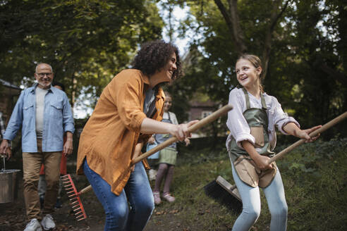 Woman and girl pretending to ride on broom sticks at community garden - HAPF03673