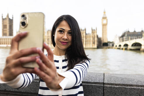 Smiling woman taking selfie with Big Ben and Houses of Parliament through smart phone in London city - WPEF07956