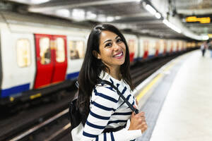 Happy woman with backpack standing in front of subway at station - WPEF07966