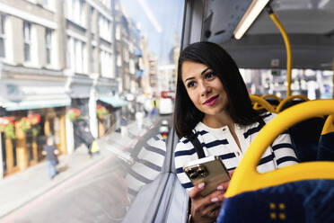 Smiling woman holding smart phone and looking through window in bus - WPEF08005