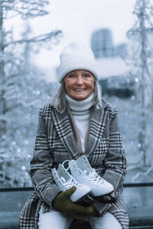 Portrait of happy senior woman in winter at an outdoor ice skating rink. Elderly woman holding brand new ice skates. - HPIF32541